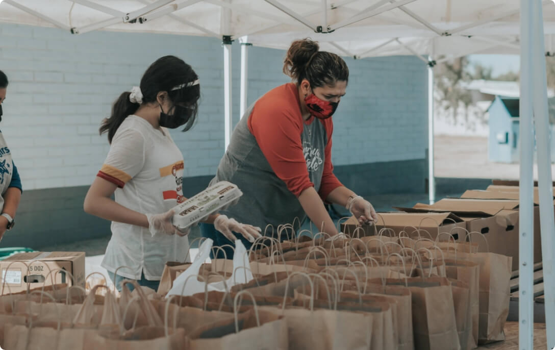 Two women volunteers packing food into paper bags under a volunteer tent