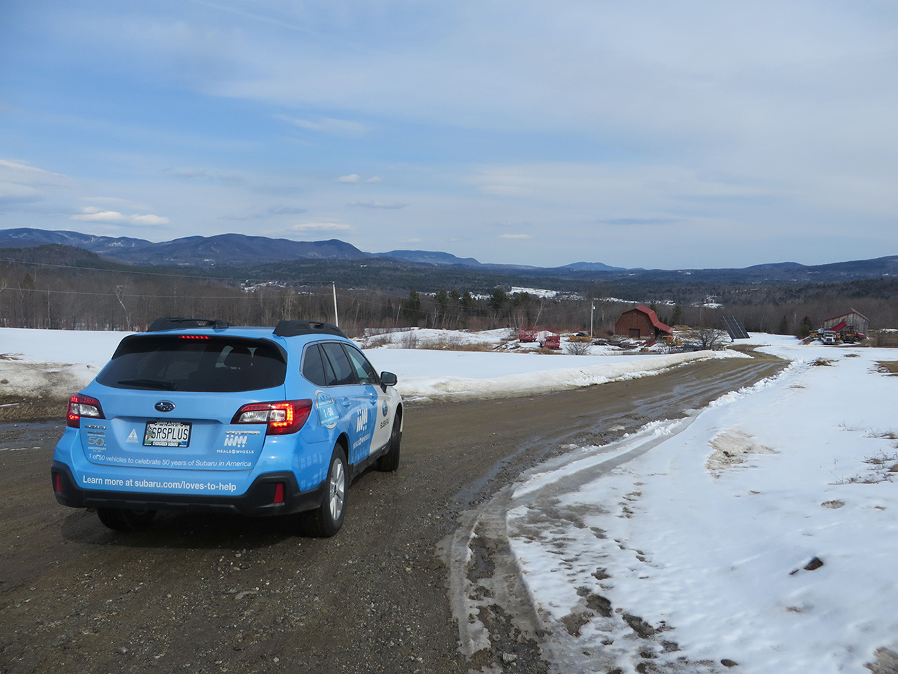 A blue Subaru with Meals on Wheels logos is stopped on a dirt road. There is snow on the ground and the mountains of Western Maine are visible in the distance.